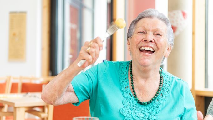 Lachende Seniorin beim Mittagstisch hält Kartoffel auf Gabel
